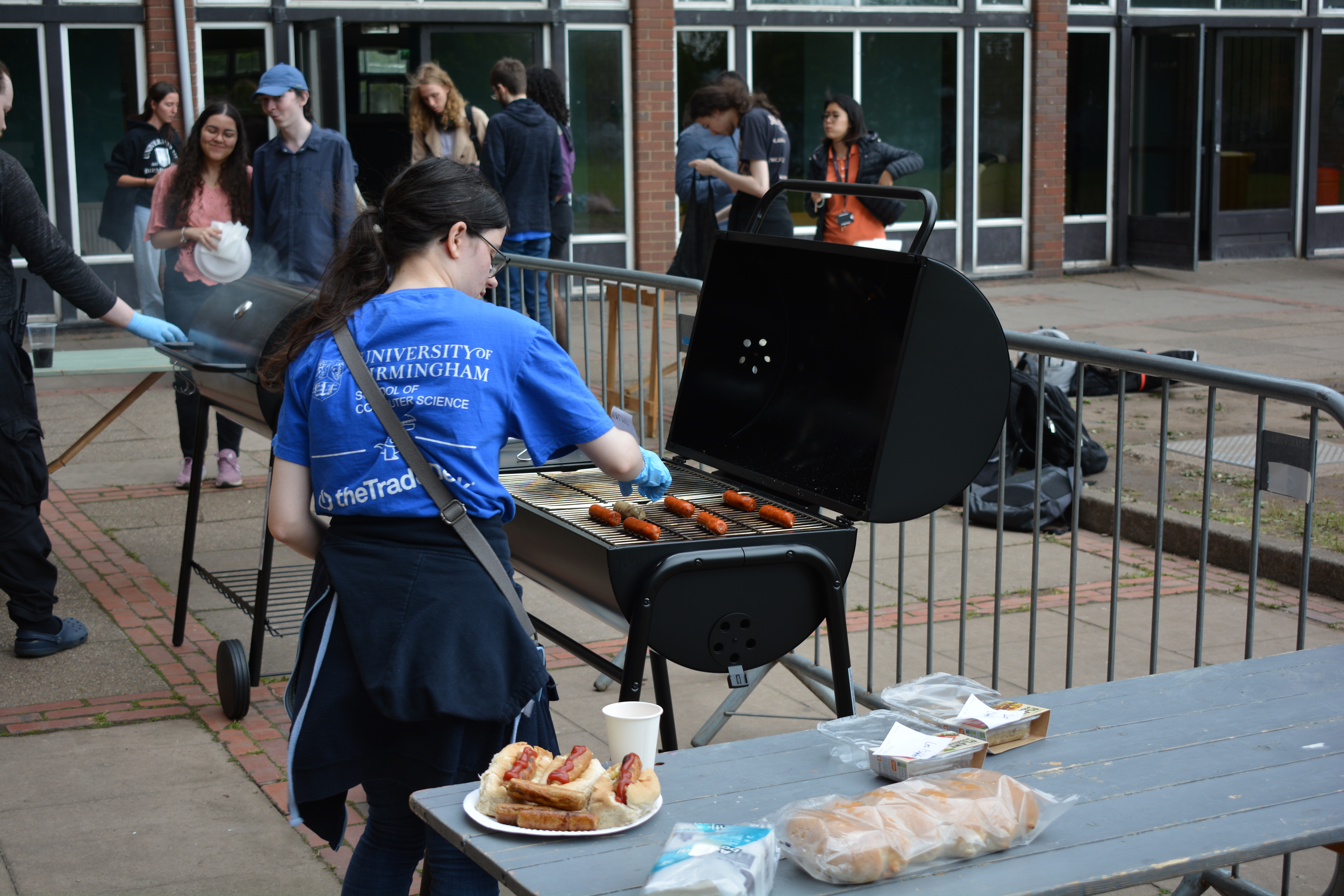 A photo of the BBQ event, with a BBQ in the foreground and a group of people in the background.